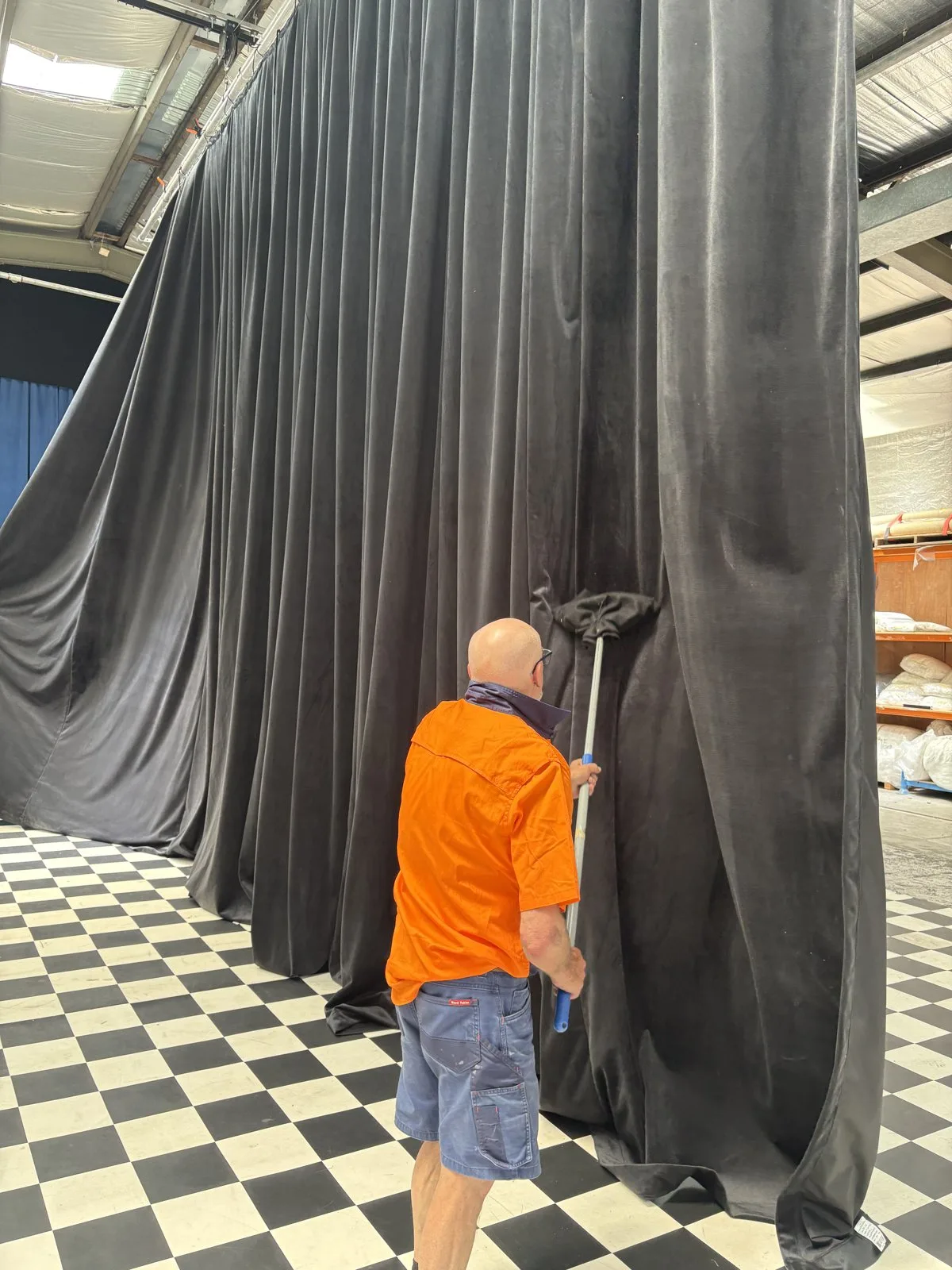 A technician cleaning a hung black velvet drape with a long-handled brush