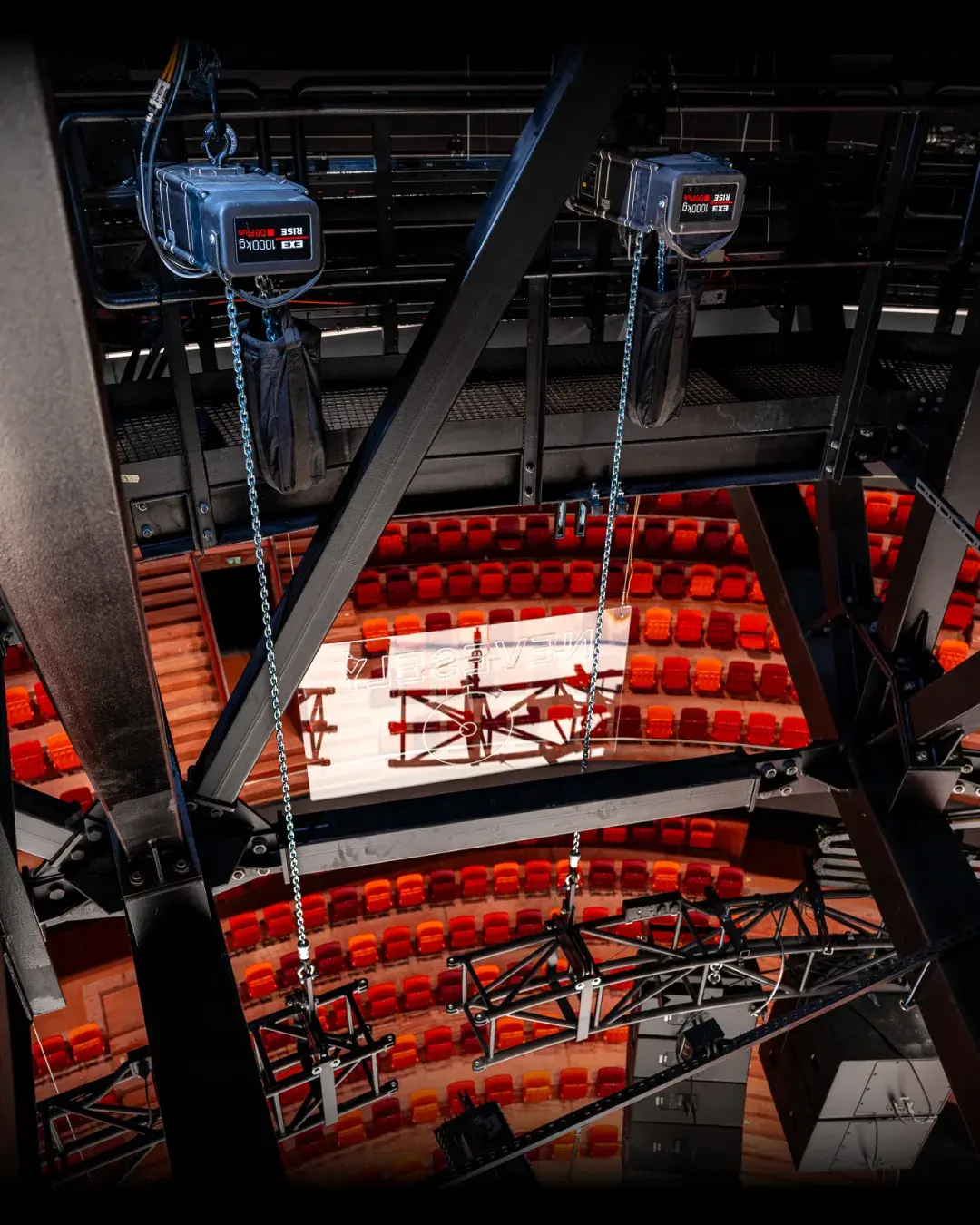 Two chain hoists flown in the overhead grid above a red-seated auditorium, chains dropping toward the stage.