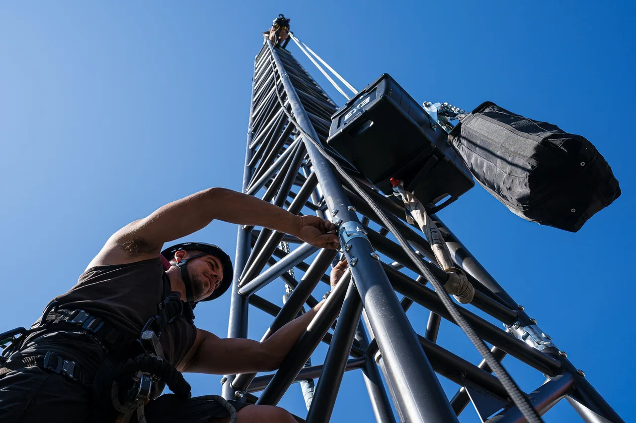 A rigger climbing an outdoor truss tower with an EXE chain hoist and chain bag overhead