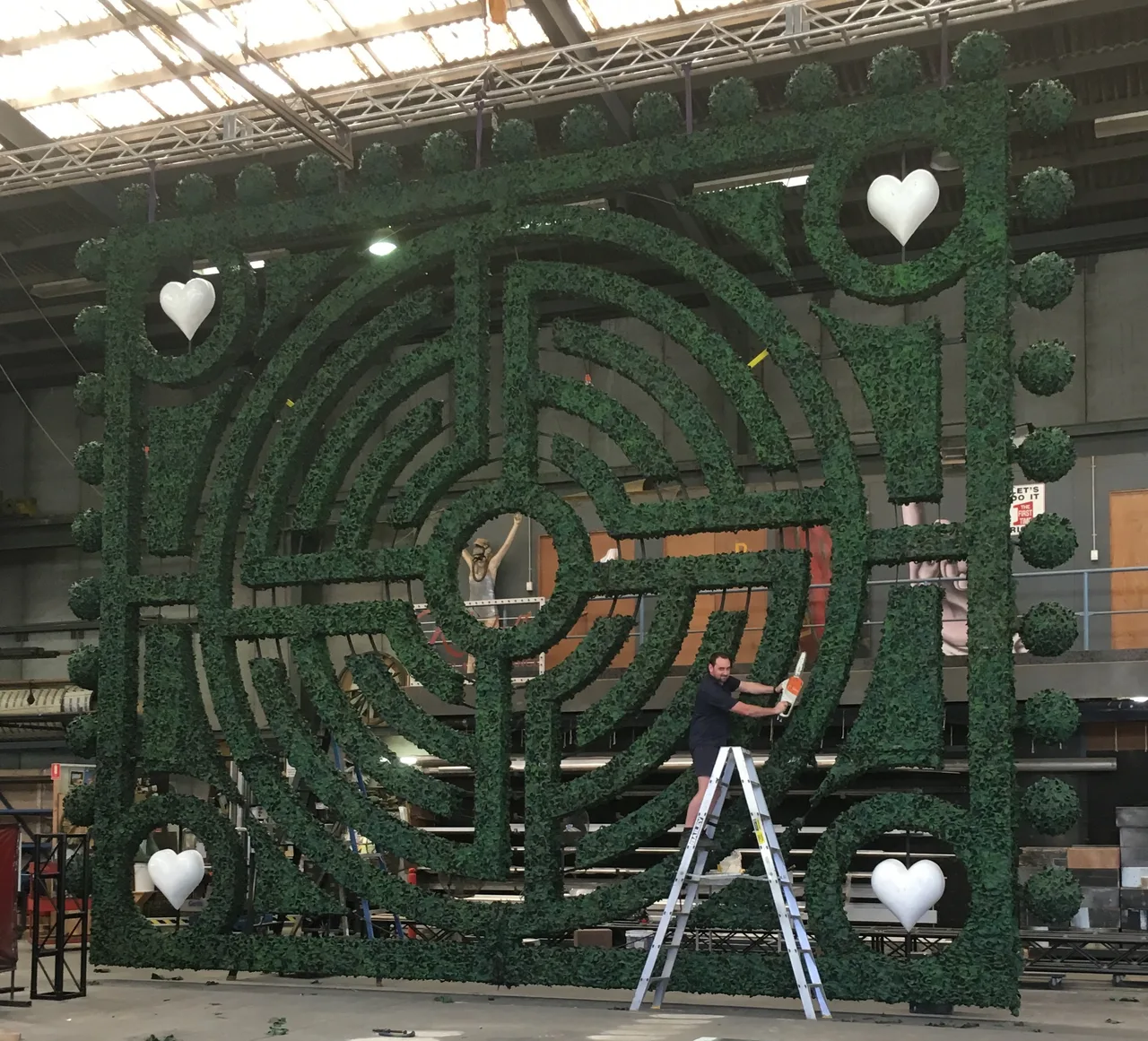 A scenic artist trimming a giant faux hedge-maze wall with white hearts