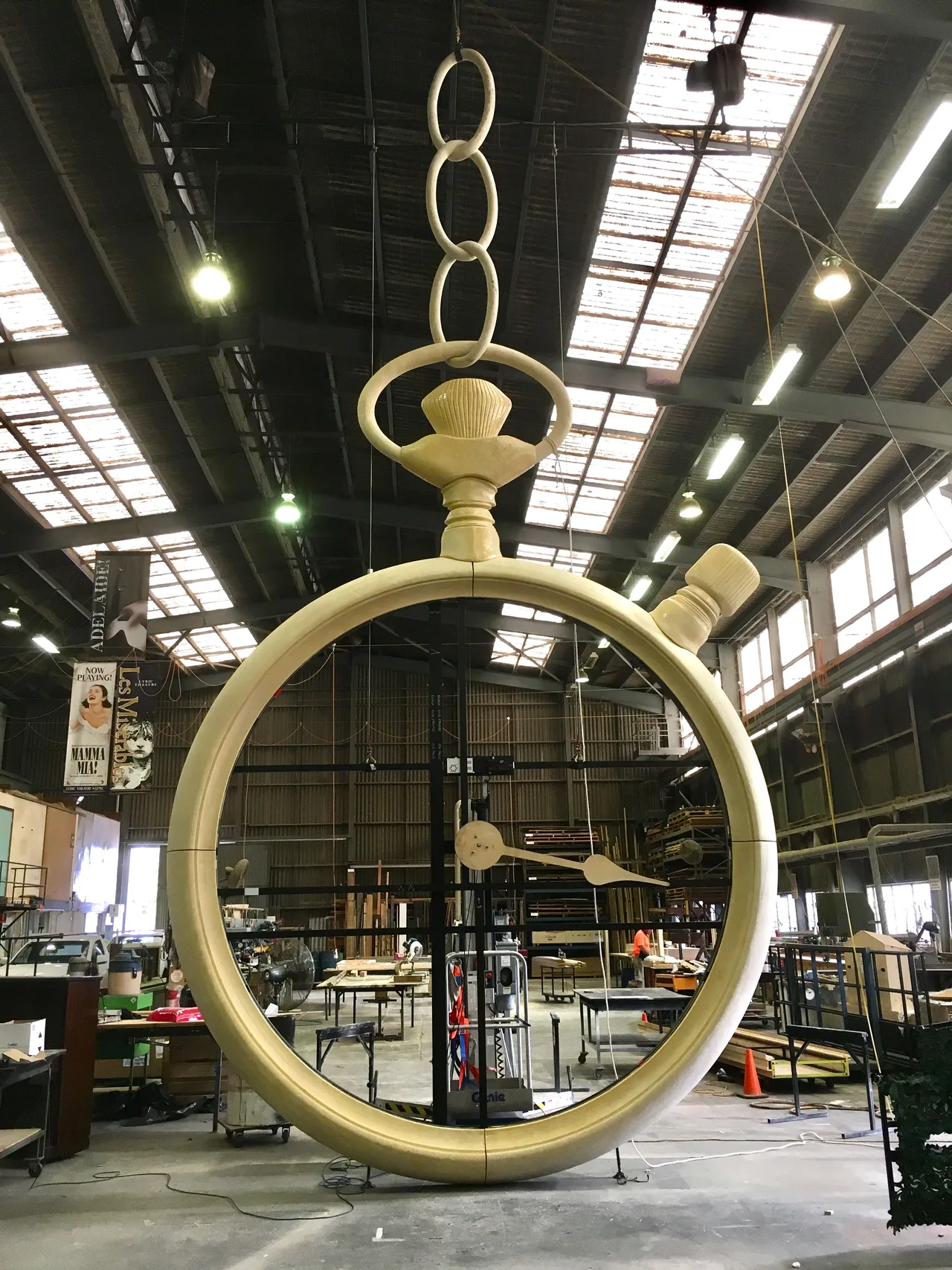 A giant sculpted pocket-watch set piece suspended from rigging in the scenic workshop